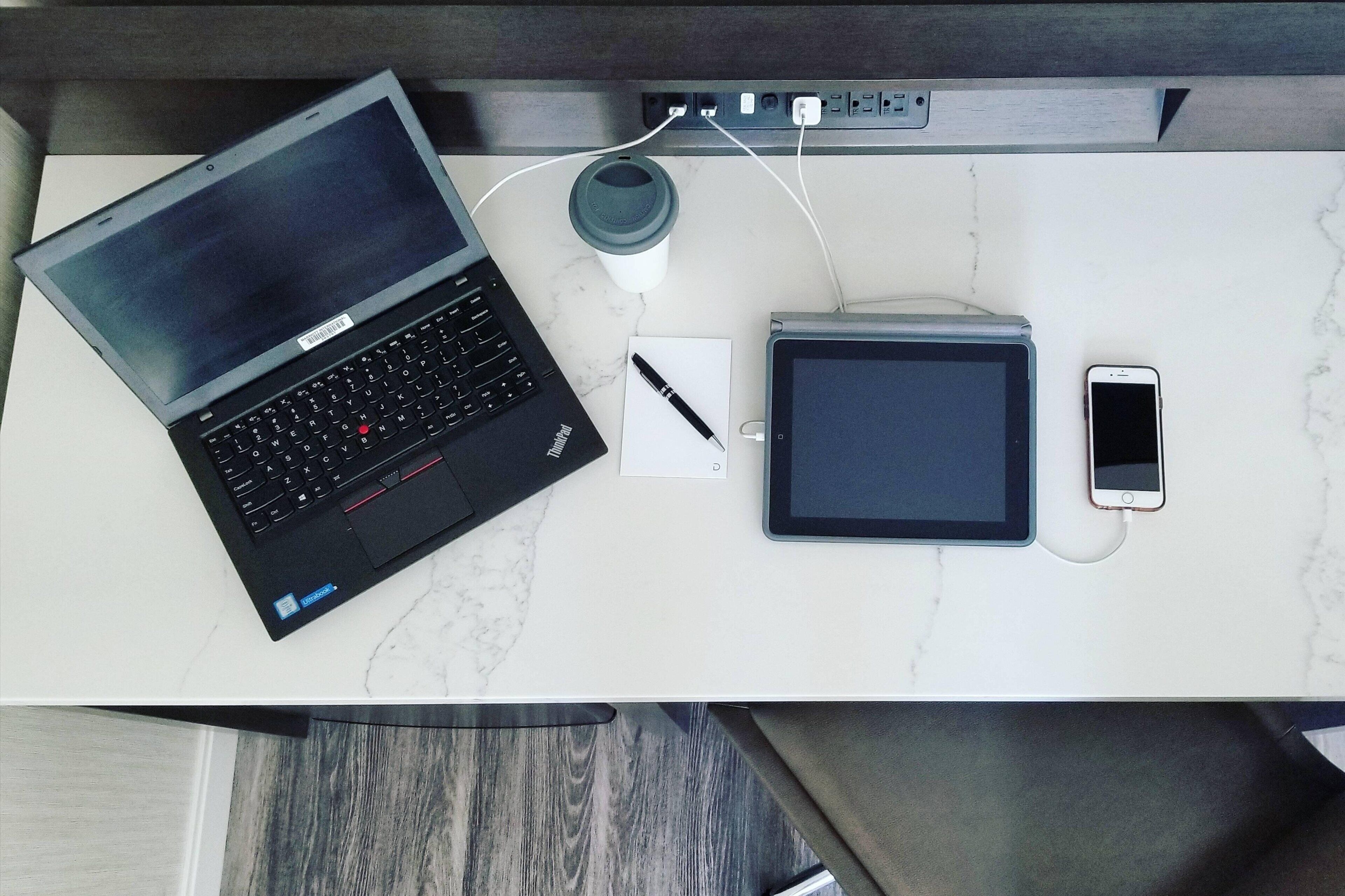 Een laptop, tablet, smartphone, koffiekop, pen en notitieboekje op een marmeren aanrecht.