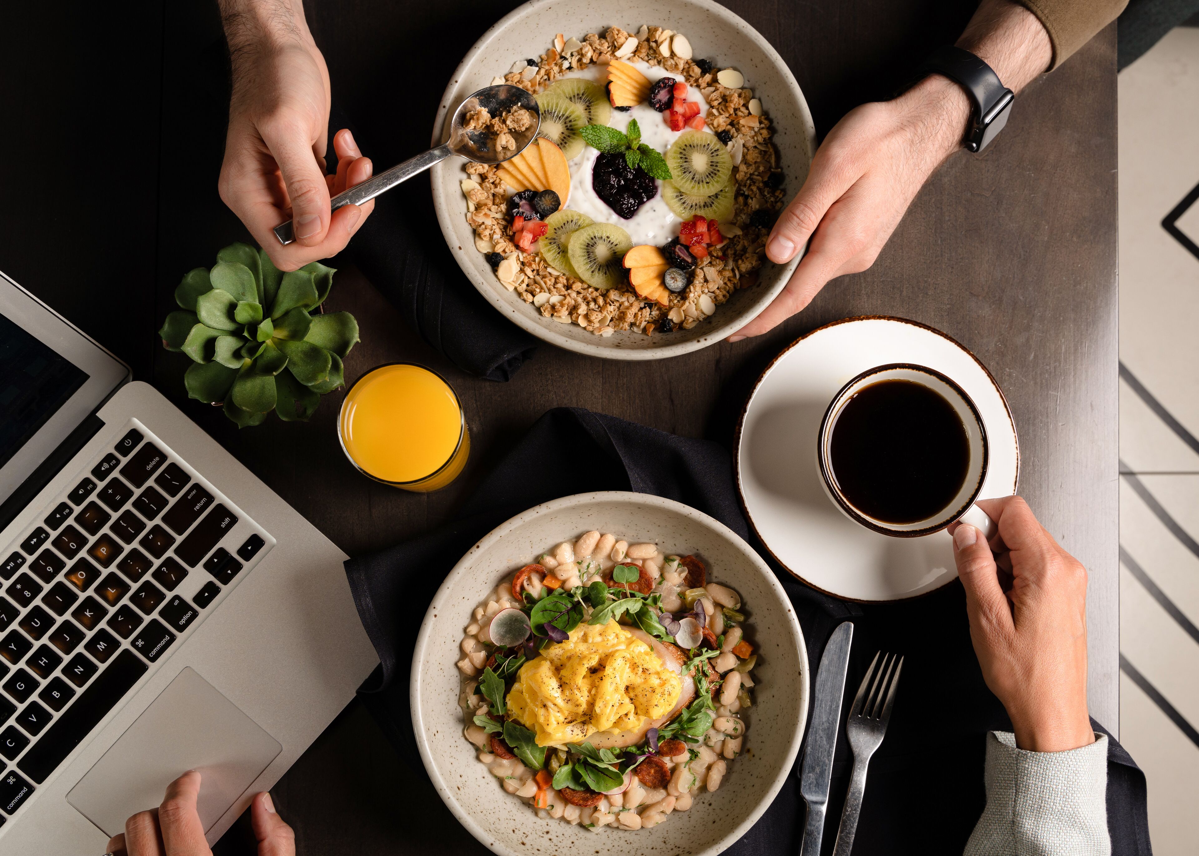 Een ontbijtsetting met een laptop, een glas sinaasappelsap, een kop koffie en twee kommen eten.