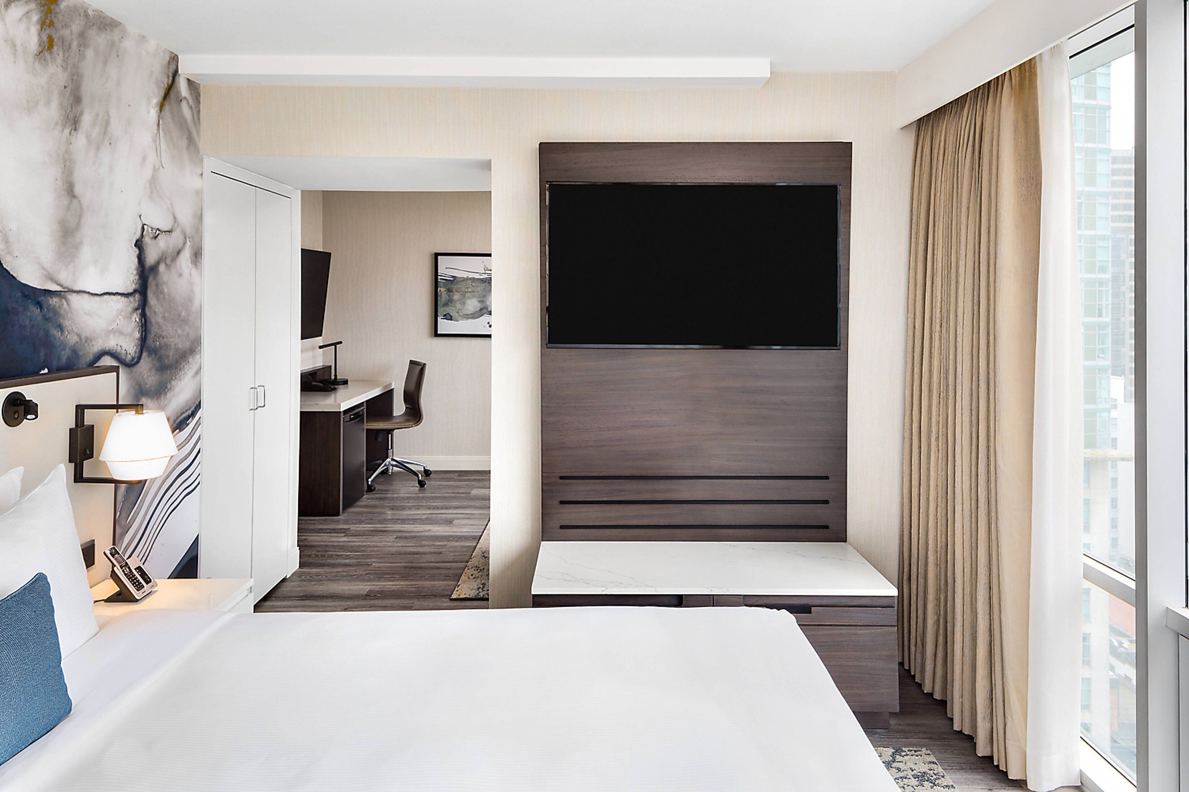 A modern hotel room with a large bed, a desk, a flat-screen TV, and a view of the city.
