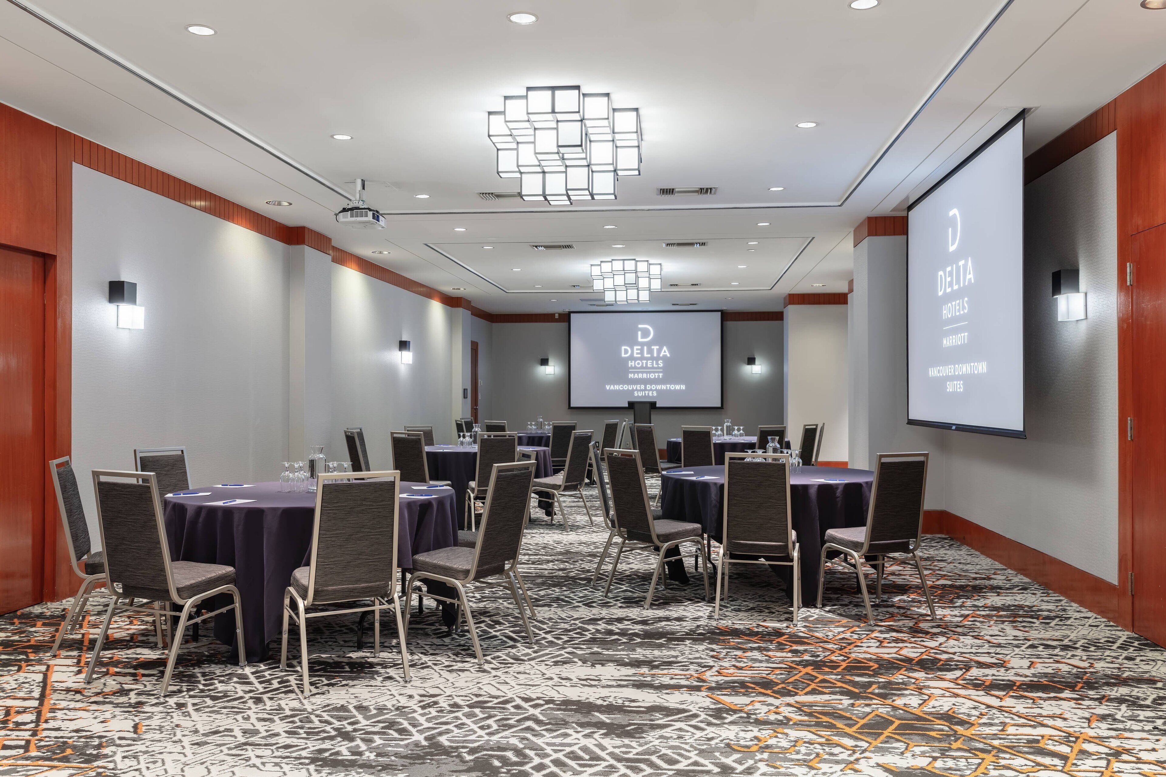 Ein Konferenzraum mit Tischen, Stühlen, einer Projektionsleinwand und einem großen Bildschirm mit dem Delta Hotels Logo.
