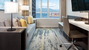 Ein Hotelzimmer mit einer Couch, einem Schreibtisch, einem Fernseher und Blick auf die Stadt und das Wasser.