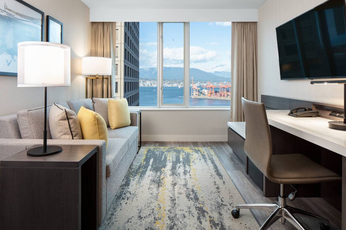 Habitación de hotel con sofá, escritorio, televisor y vistas a la ciudad y al agua.