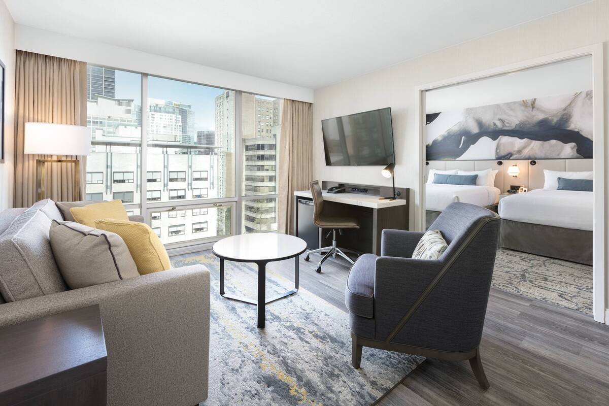 Una habitación de hotel moderna con un ventanal grande, un televisor de pantalla plana, un escritorio, un sofá cómodo y una cama con vistas a la ciudad.