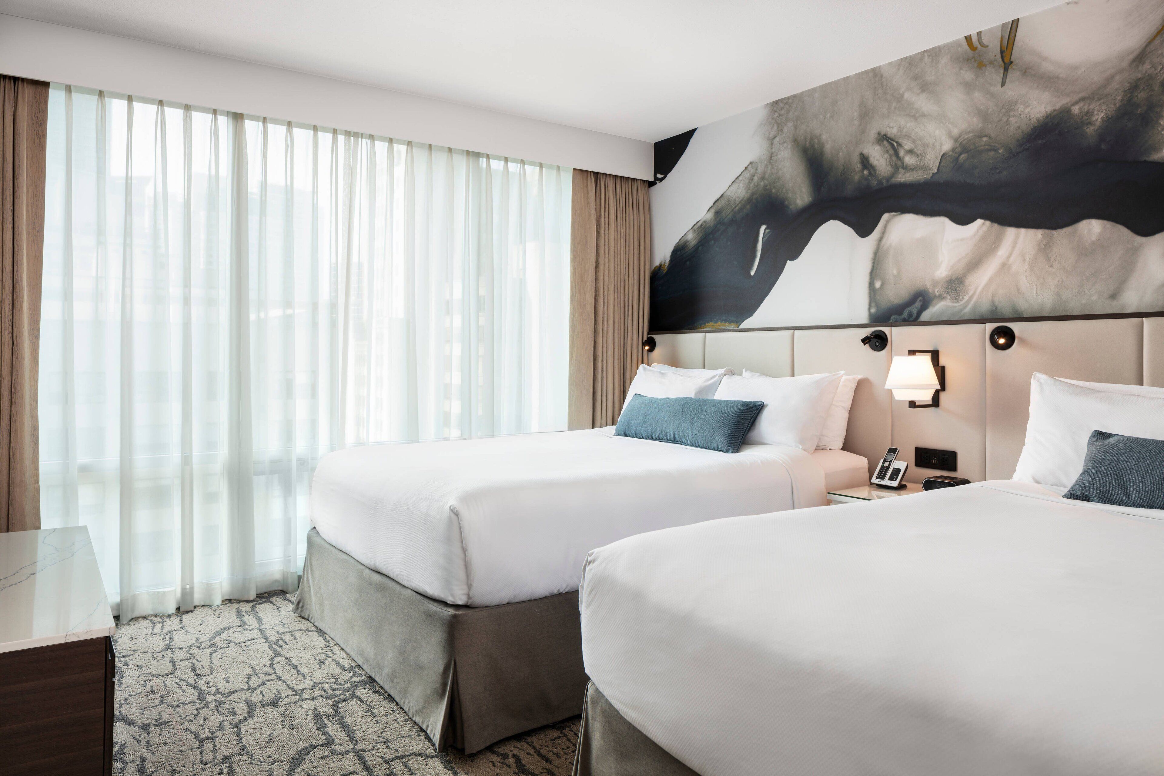 Habitación de hotel con dos camas, un cuadro abstracto grande en la pared, un escritorio y una ventana con cortinas translúcidas.