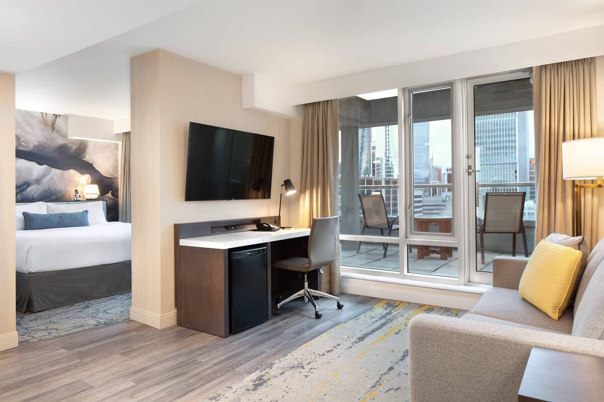 Habitación de hotel con cama, escritorio, silla y un amplio ventanal con vistas a la ciudad.