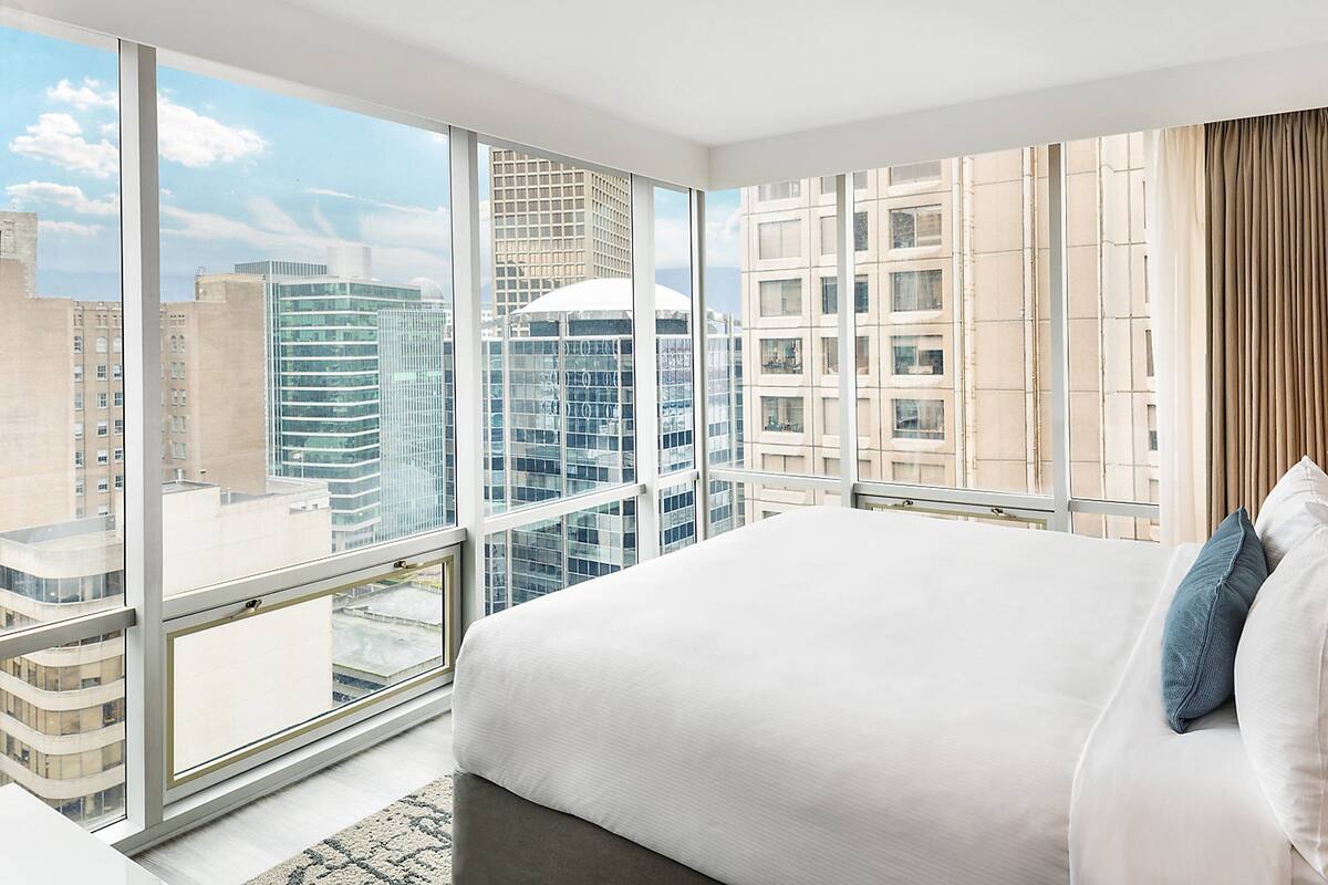 Una habitación de hotel moderna con una cama grande, vistas del paisaje urbano a través de ventanas de piso a techo y un horizonte de la ciudad al fondo.