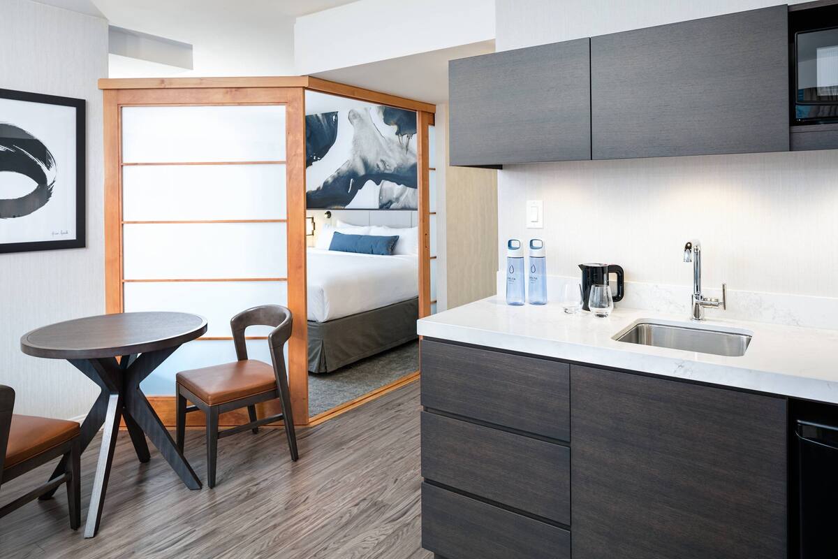 Una habitación de hotel compacta con kitchenette, una cama con almohada azul, una mesa de comedor redonda con dos sillas y un cuadro abstracto grande en la pared.