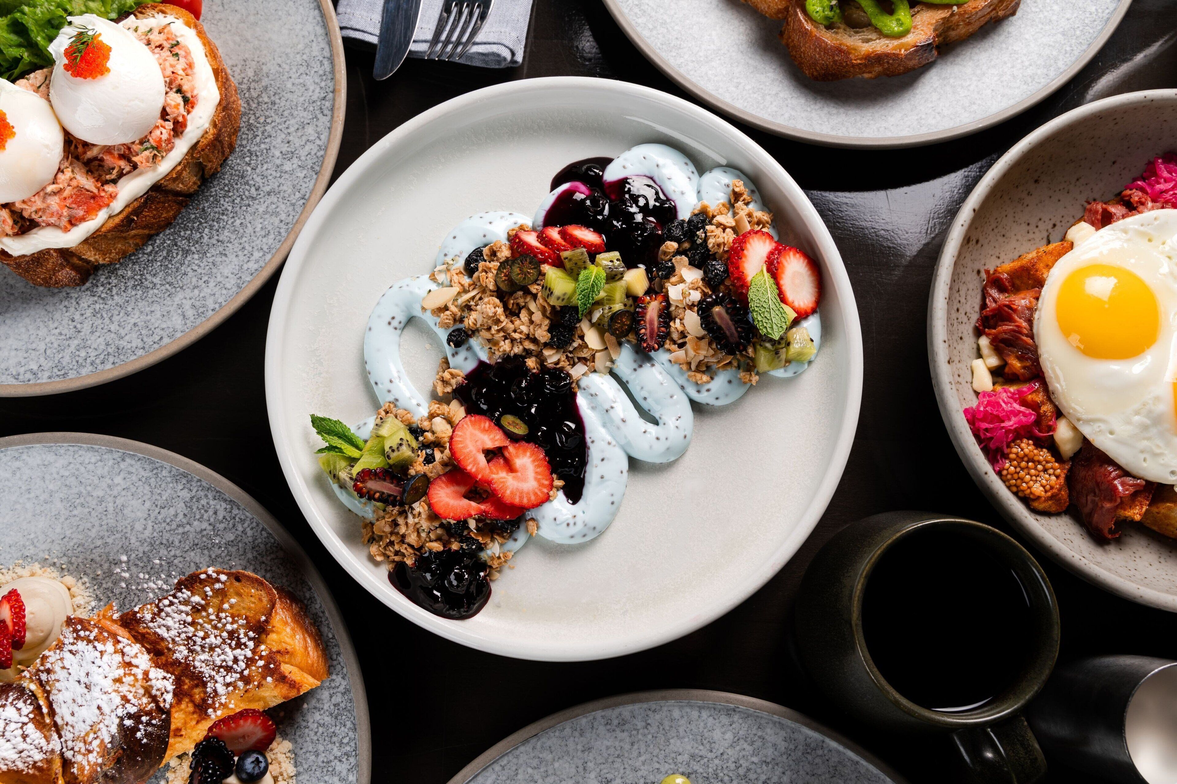 Une variété de plats pour le petit-déjeuner sont présentés sur des assiettes, notamment des toasts avec des œufs, un bol de yaourt aux fruits rouges et une assiette avec un œuf au plat et des légumes.
