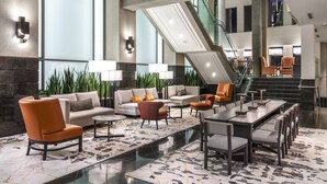 Eine großzügige Lobby mit modernem Design, ausgestattet mit einer geräumigen Sitzecke, Sofas und Sesseln, einem langen Esstisch sowie einer Treppe zu einer oberen Ebene.