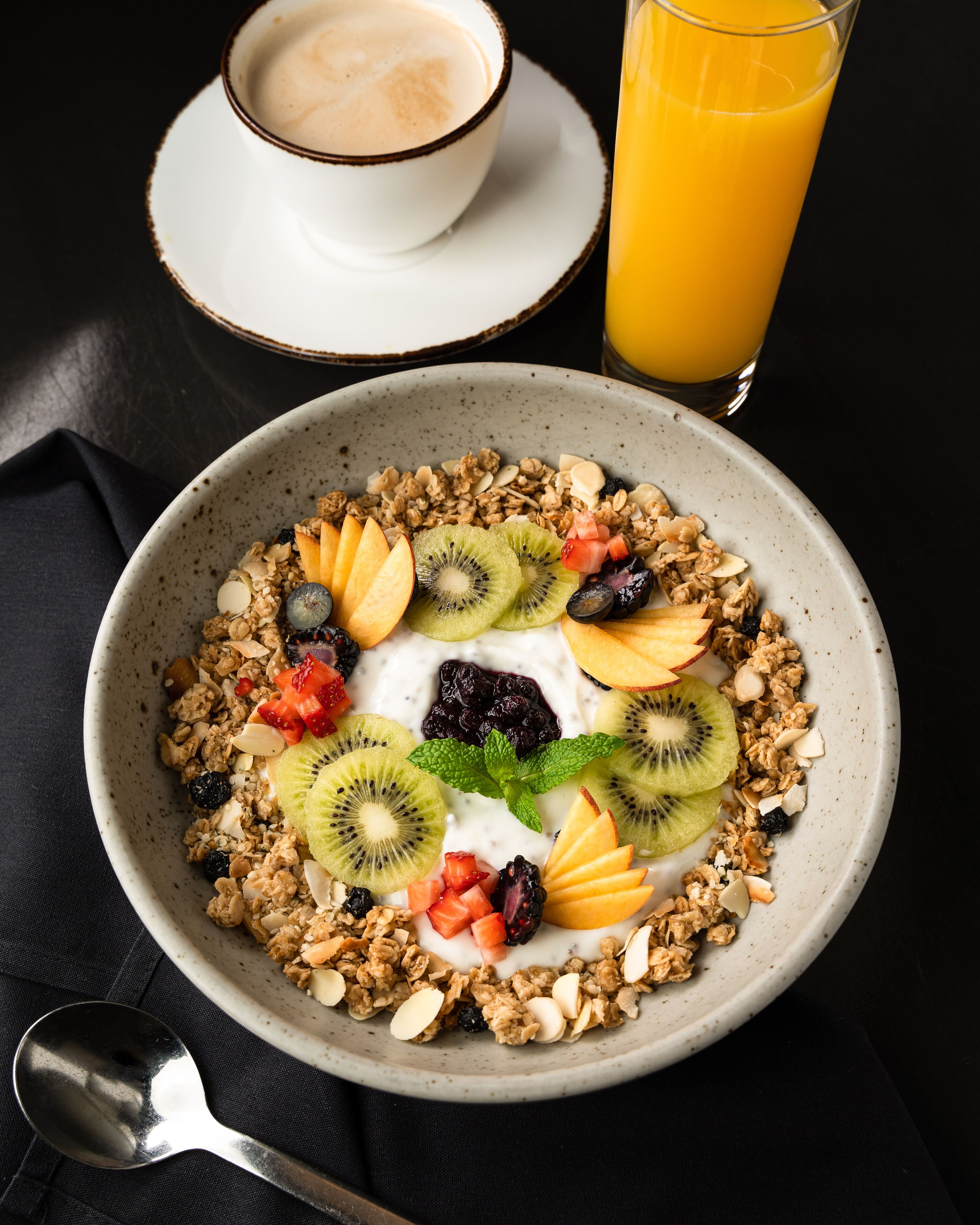 Un tazón de granola con frutas y nueces, un vaso de jugo de naranja y una taza de café sobre una mesa.