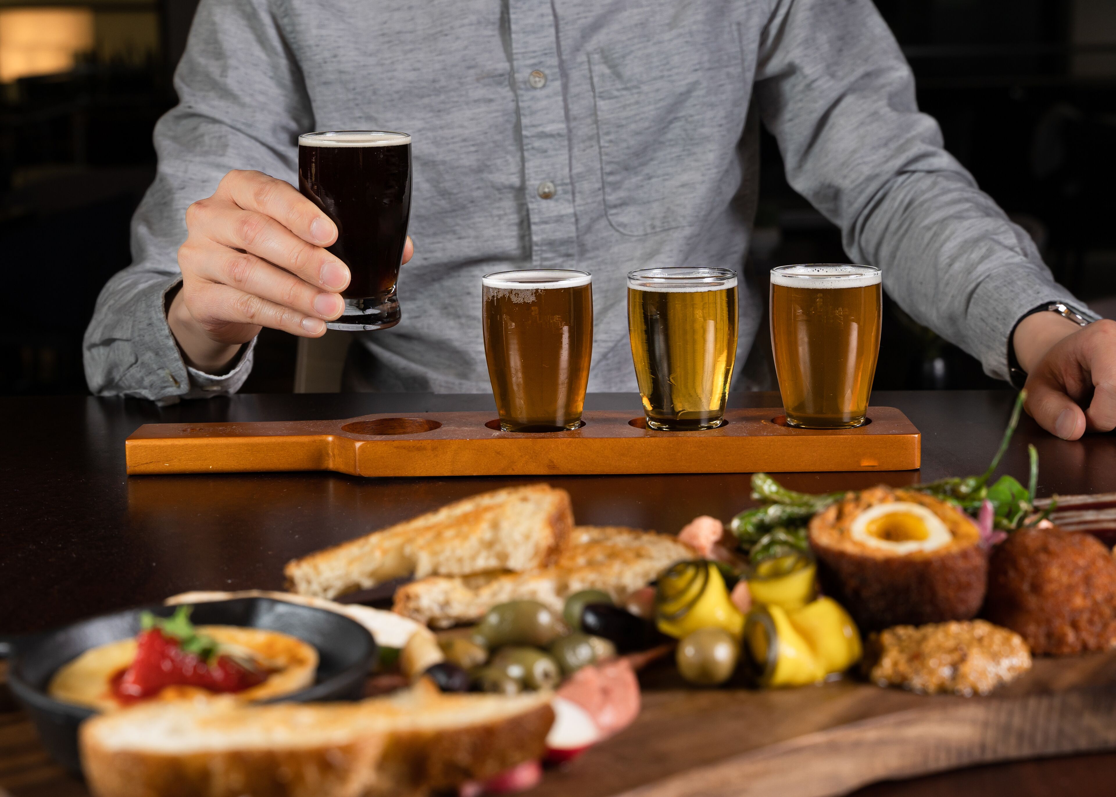 Una persona sosteniendo un vaso de cerveza oscura, con tres vasos más de cerveza en una bandeja de madera. Hay una variedad de comida en la mesa, incluyendo pan, aceitunas y pepinillos.
