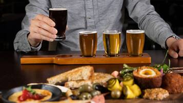 Una persona sosteniendo un vaso de cerveza oscura, con tres vasos más de cerveza en una bandeja de madera. Hay una variedad de comida en la mesa, incluyendo pan, aceitunas y pepinillos.