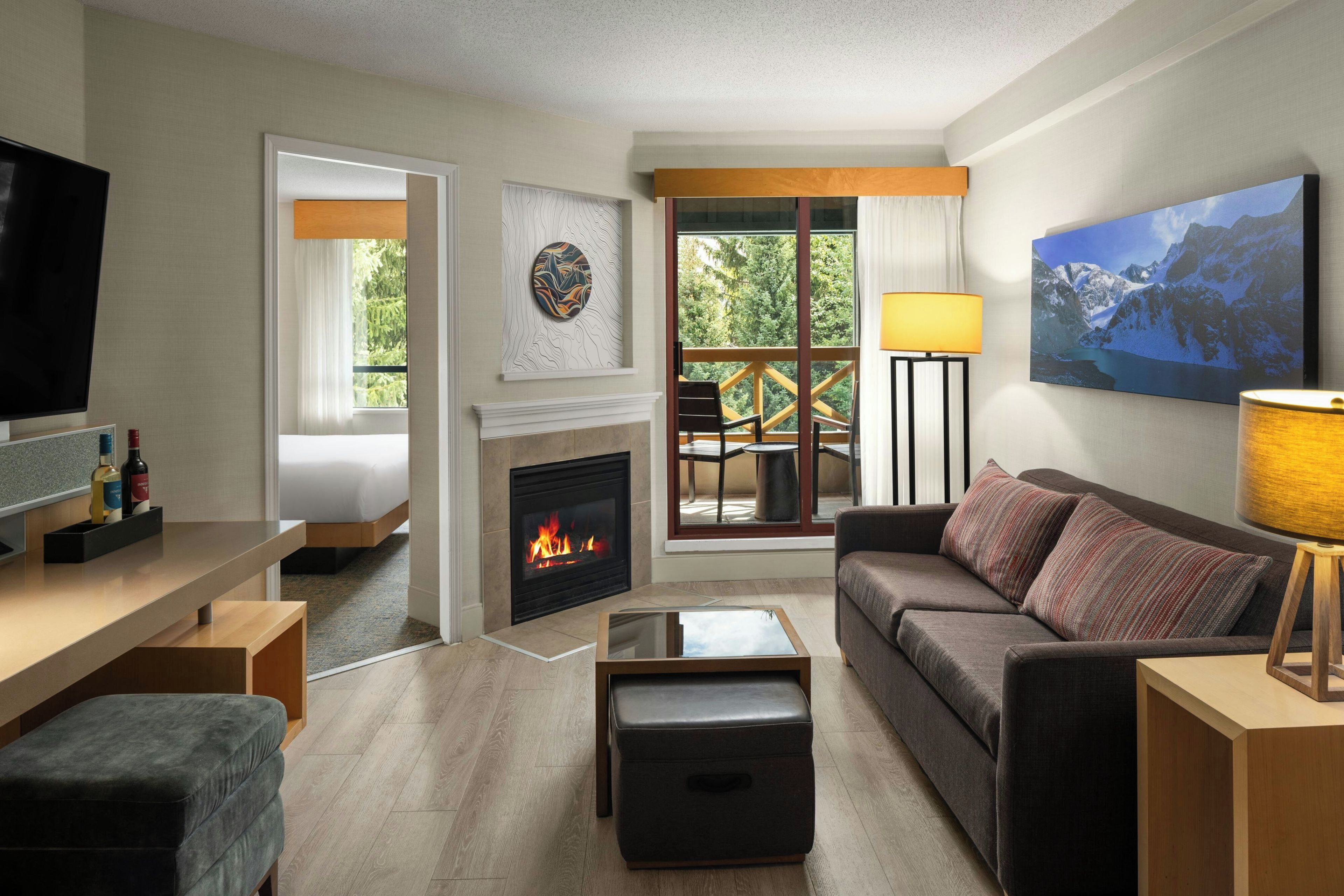 A modern living room with a fireplace, a sofa, a TV, and a balcony with a view of trees.