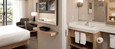 Habitación de hotel con cama, zona de estar, baño con lavamanos y espejo, y vista al exterior.