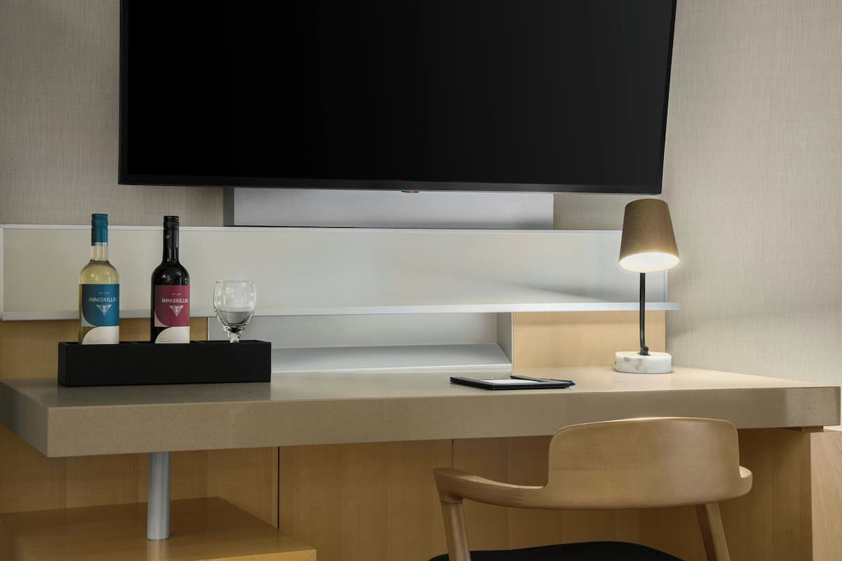 A modern hotel room with a flat-screen TV mounted on the wall, a desk with a lamp, a chair, and a tray with two bottles and a glass.