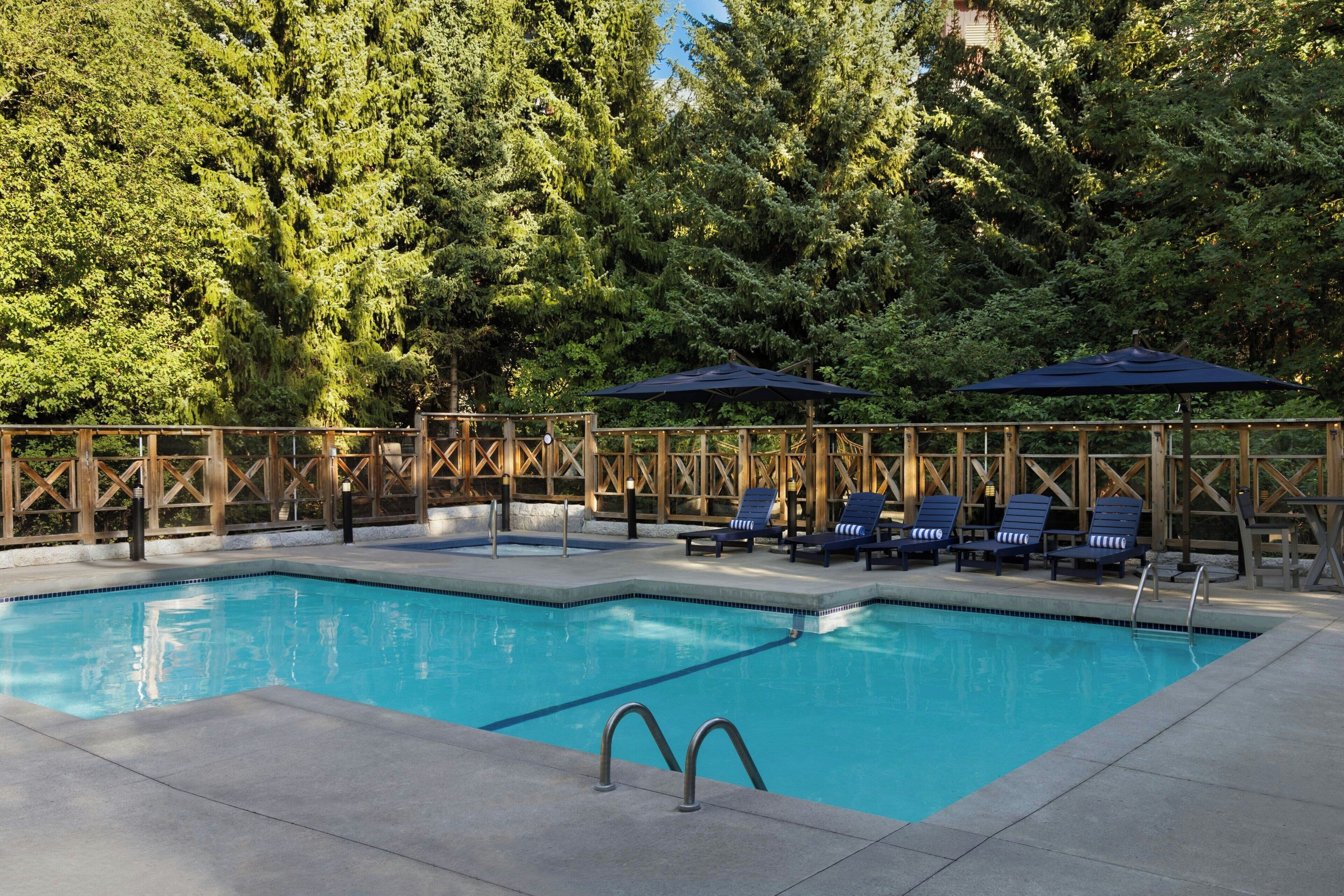 A rectangular swimming pool with a wooden deck surrounding it, equipped with lounge chairs and umbrellas.