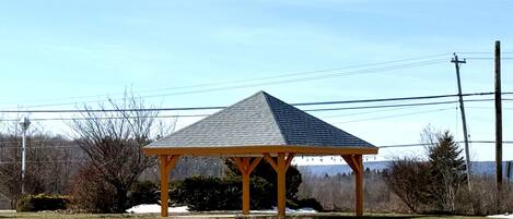 A wooden gazebo with a sloped roof in a grassy area.
