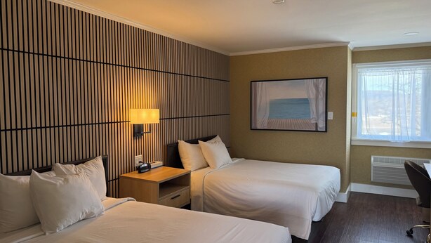 Ein Hotelzimmer mit zwei Betten, einem Nachttisch mit Lampe, einem Strandbild, einem Fenster mit Vorhängen und einem Schreibtisch mit Stuhl.