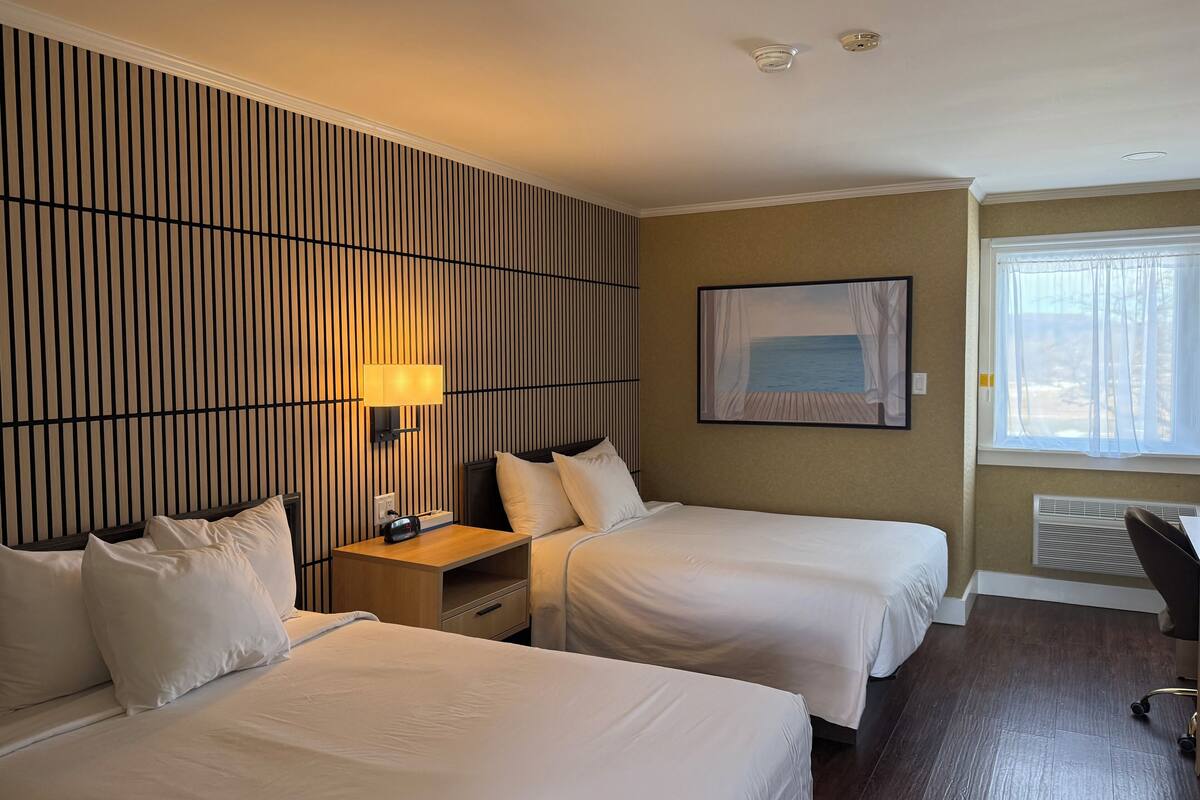 Habitación de hotel con dos camas, una mesita de noche con lámpara, un cuadro de playa, una ventana con cortinas y un escritorio con silla.