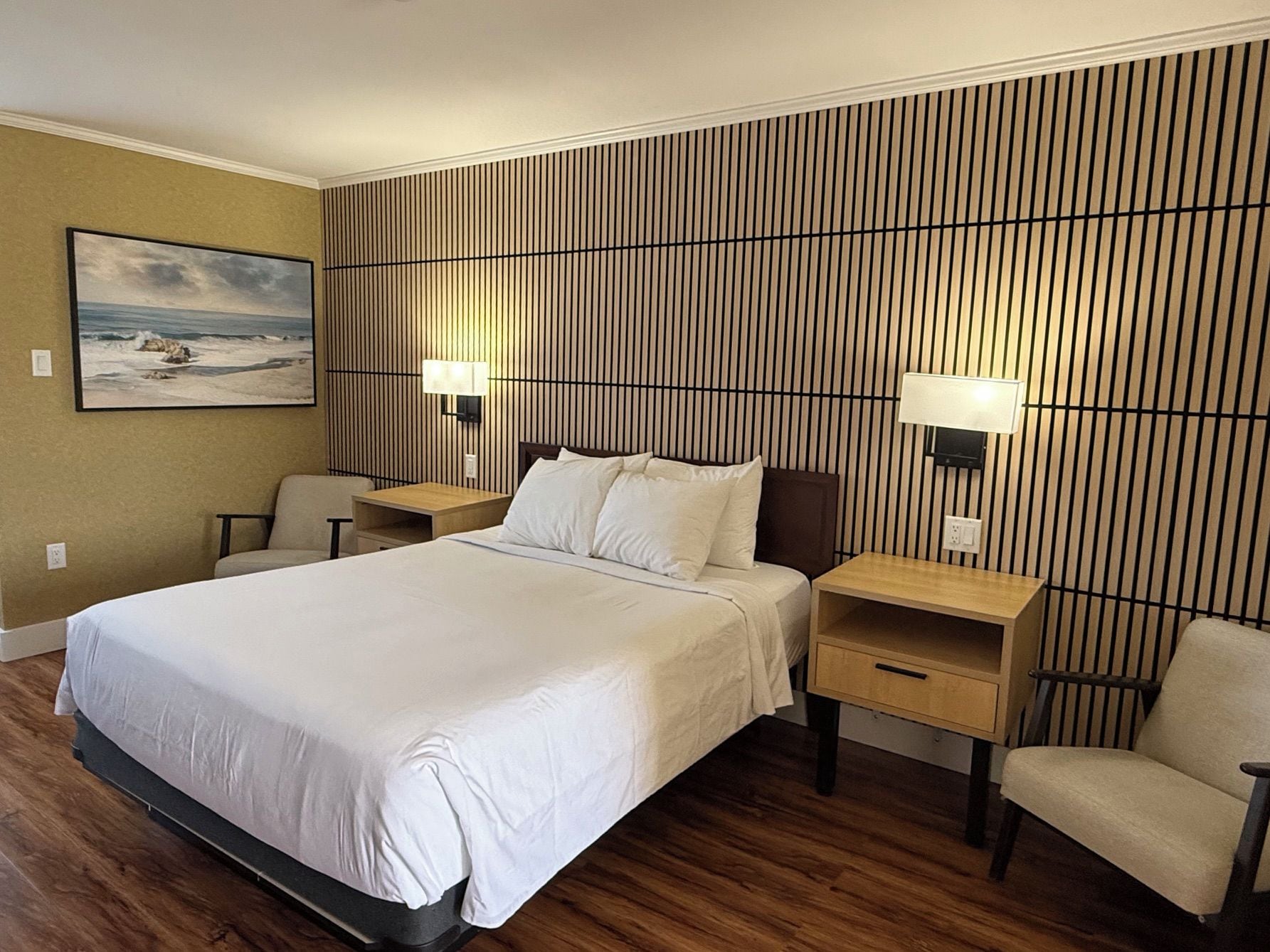 A hotel room with a large bed, two bedside tables, a chair, a wall-mounted TV, and a painting of a beach scene.
