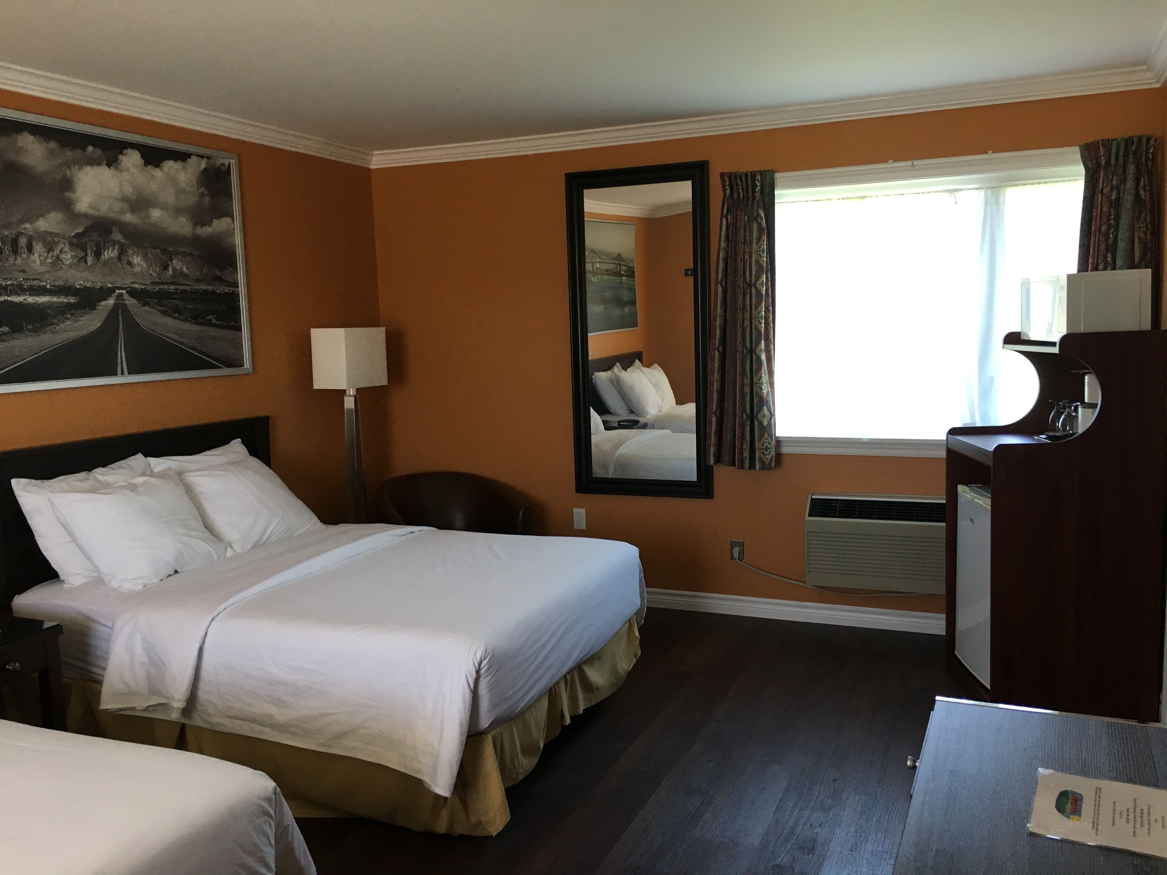 A hotel room with a bed, a desk, a chair, a lamp, a mirror, and a window with curtains.