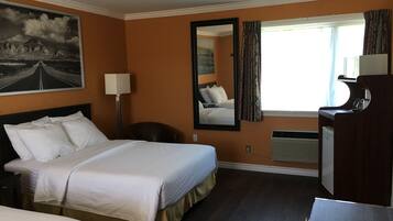 A hotel room with a bed, a desk, a chair, a lamp, a mirror, and a window with curtains.