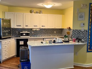 A kitchen with white cabinets labeled for glasses, cups, plates, and bowls, a microwave, a coffee maker, and a trash bin. - Annapolis Royal Inn & Suites (Annapolis Royal)