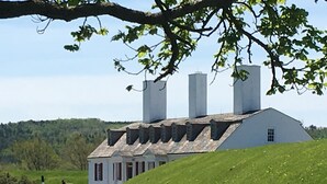 Ein weißes Gebäude mit geneigtem Dach und drei hohen Schornsteinen, gelegen auf einem grasbewachsenen Hügel mit bewaldeter Landschaft im Hintergrund.