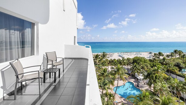 A balcony with two chairs and a small table, overlooking a pool and beach area with palm trees and a clear blue sky. - Hotel Riu Plaza Miami Beach (Miami Beach)