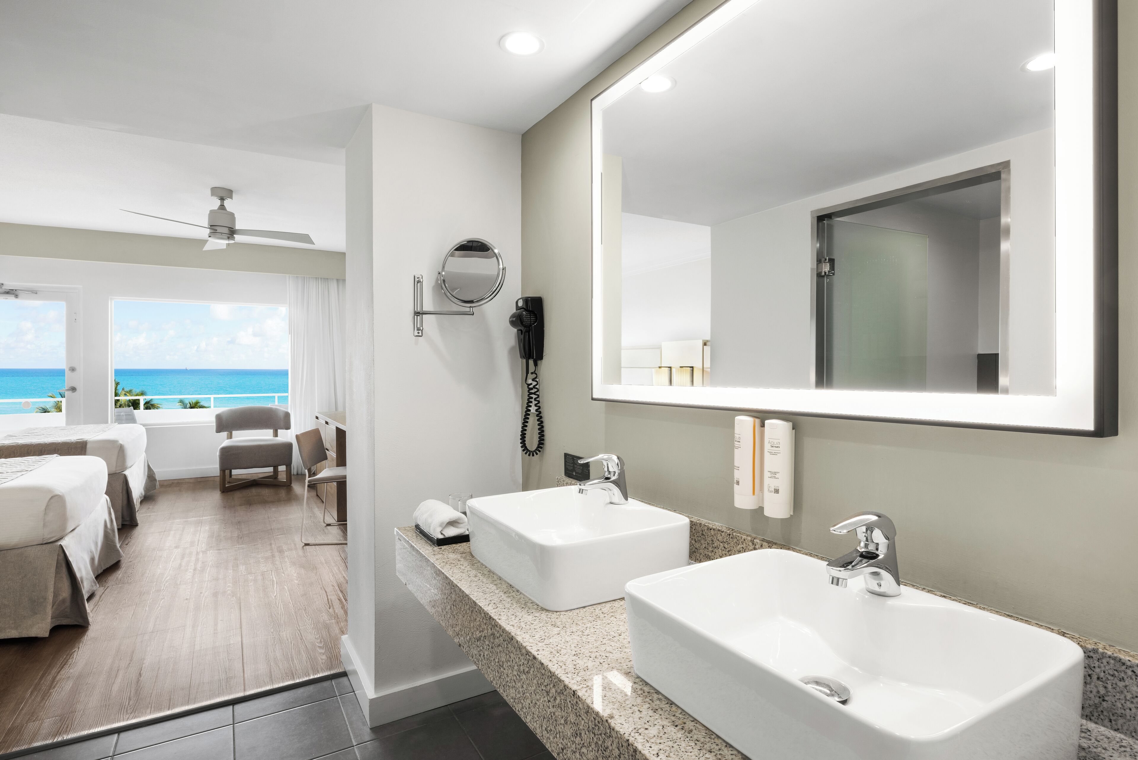 A hotel room with two large white sinks, a ceiling fan, a round mirror, a hairdryer, and a view of the ocean.