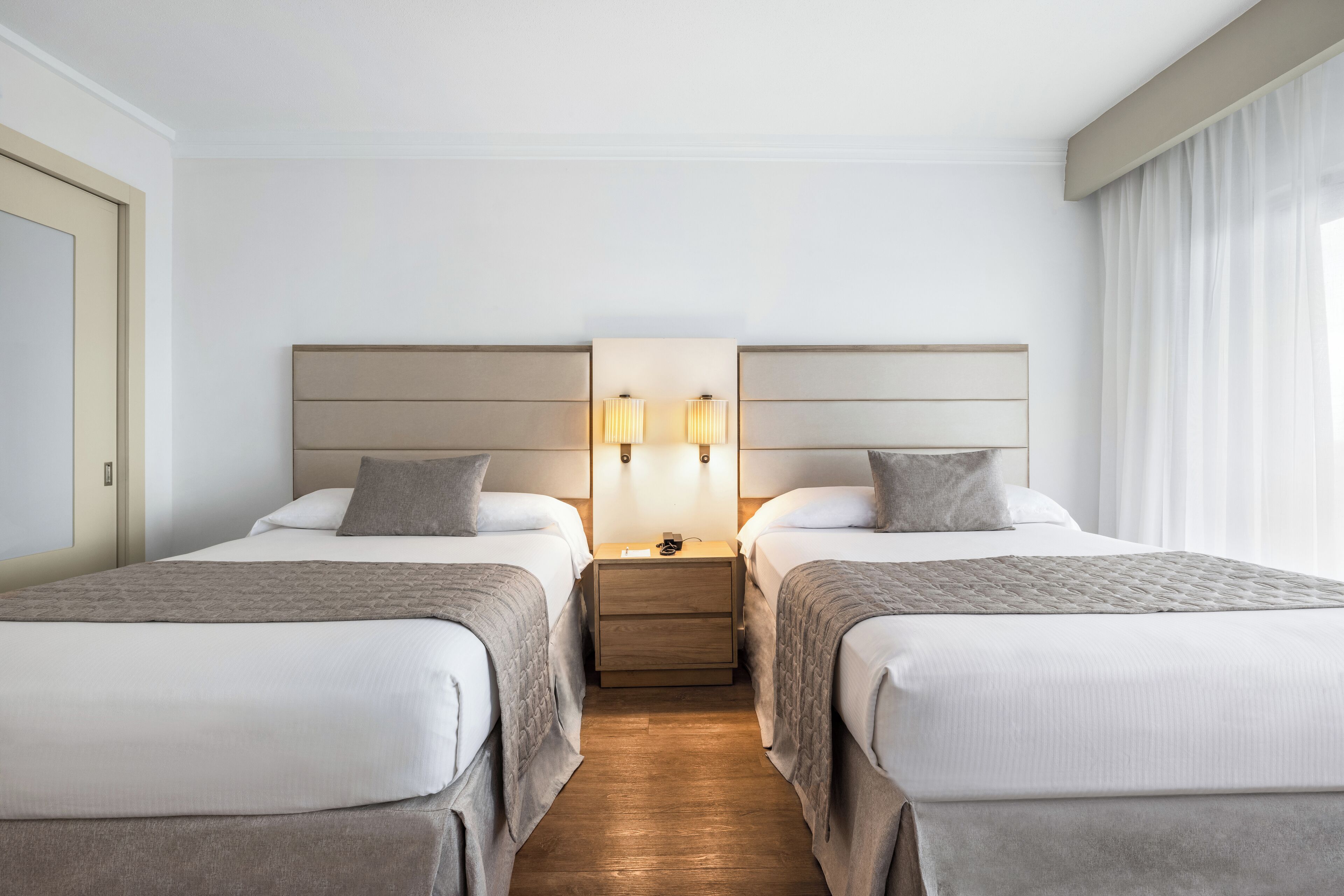 A hotel room with two beds, a nightstand with lamps, and a window with curtains.