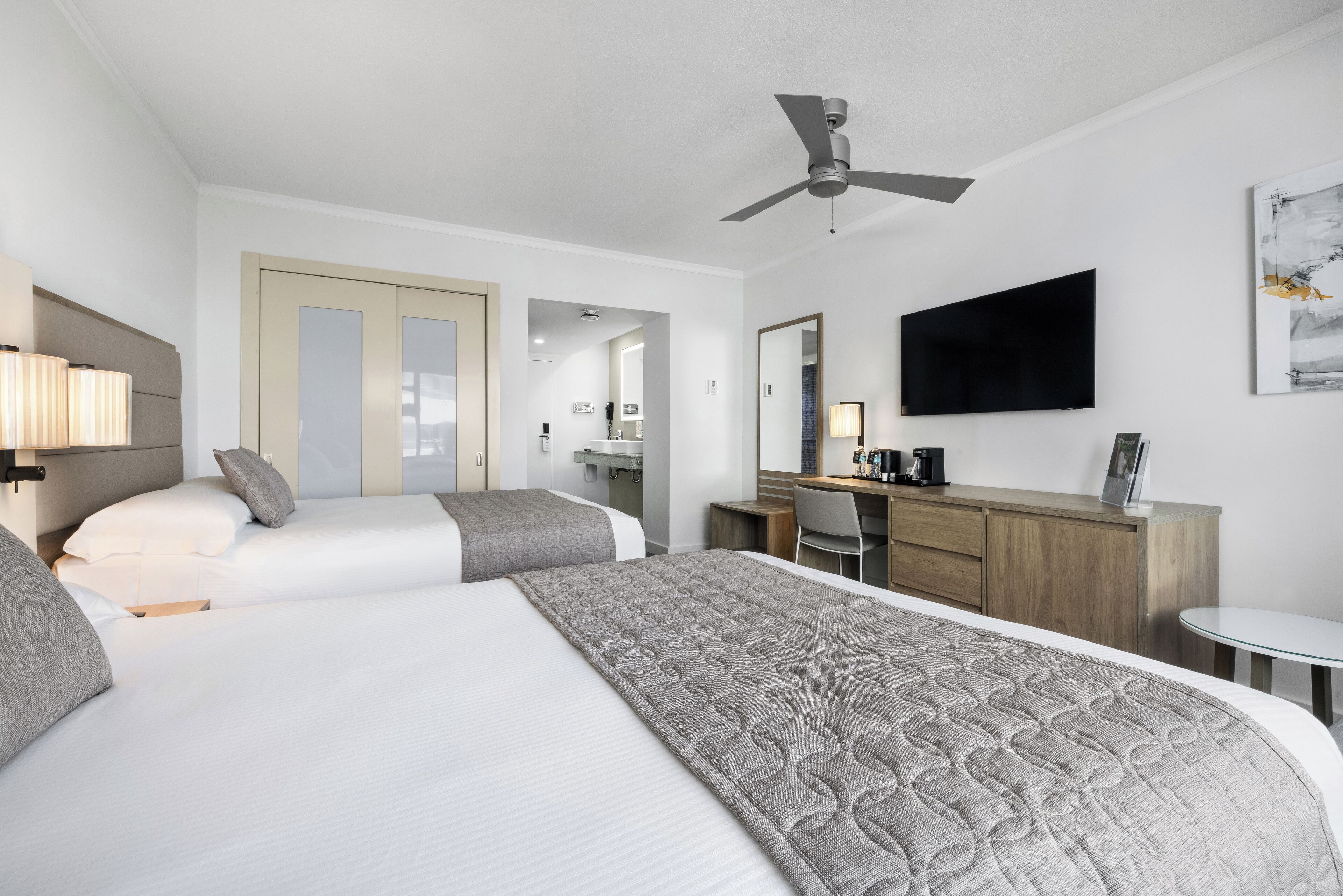 A hotel room with two beds, a desk, a chair, a TV, and a ceiling fan.