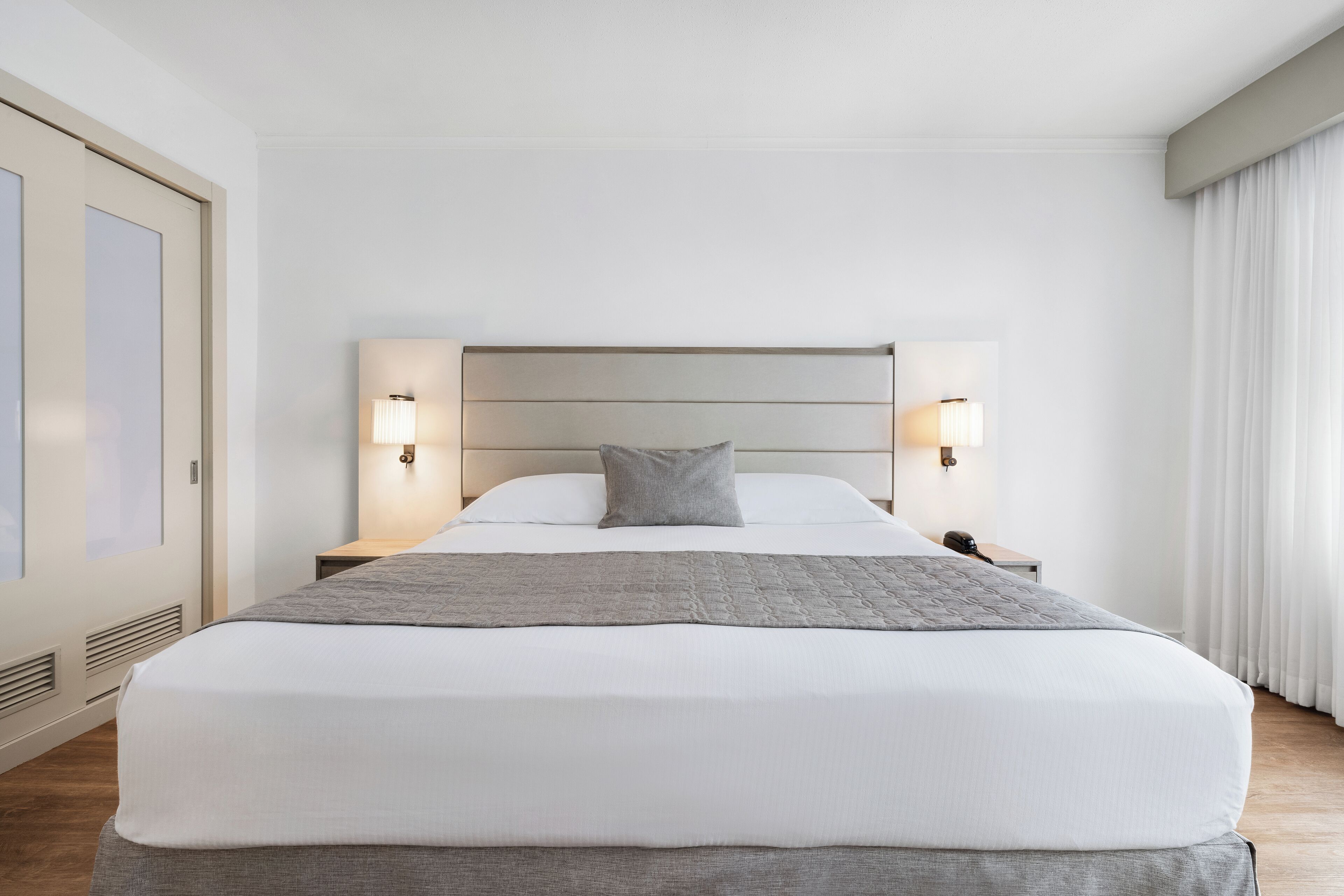 A modern hotel room with a large bed, two bedside lamps, a nightstand, and a view of the outdoors through a window with curtains.