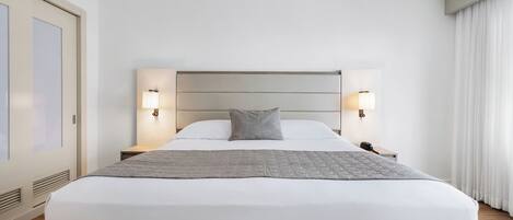 A modern hotel room with a large bed, two bedside lamps, a nightstand, and a view of the outdoors through a window with curtains.