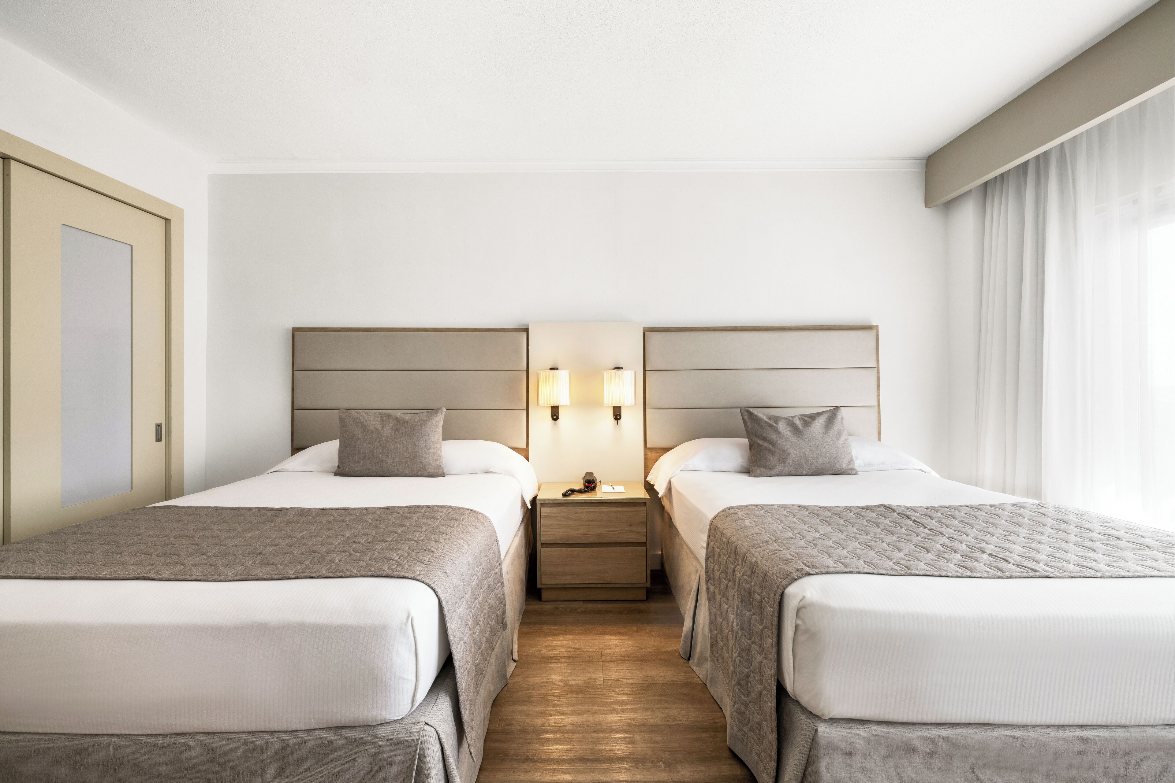 A hotel room with two beds, a nightstand with lamps, and a wooden floor.