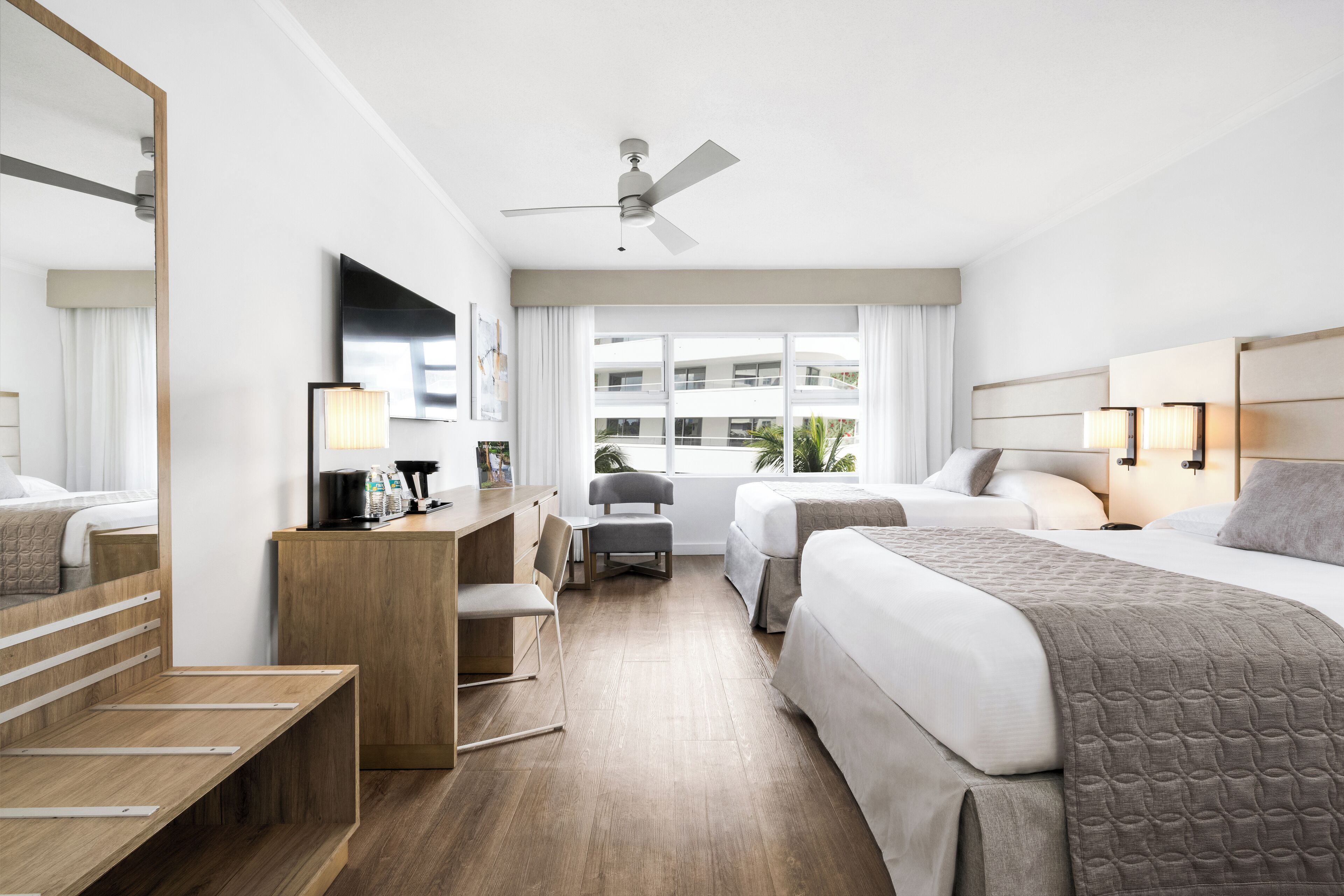 Habitación de hotel con cama, escritorio, silla, ventilador de techo y ventana con vista a edificios.