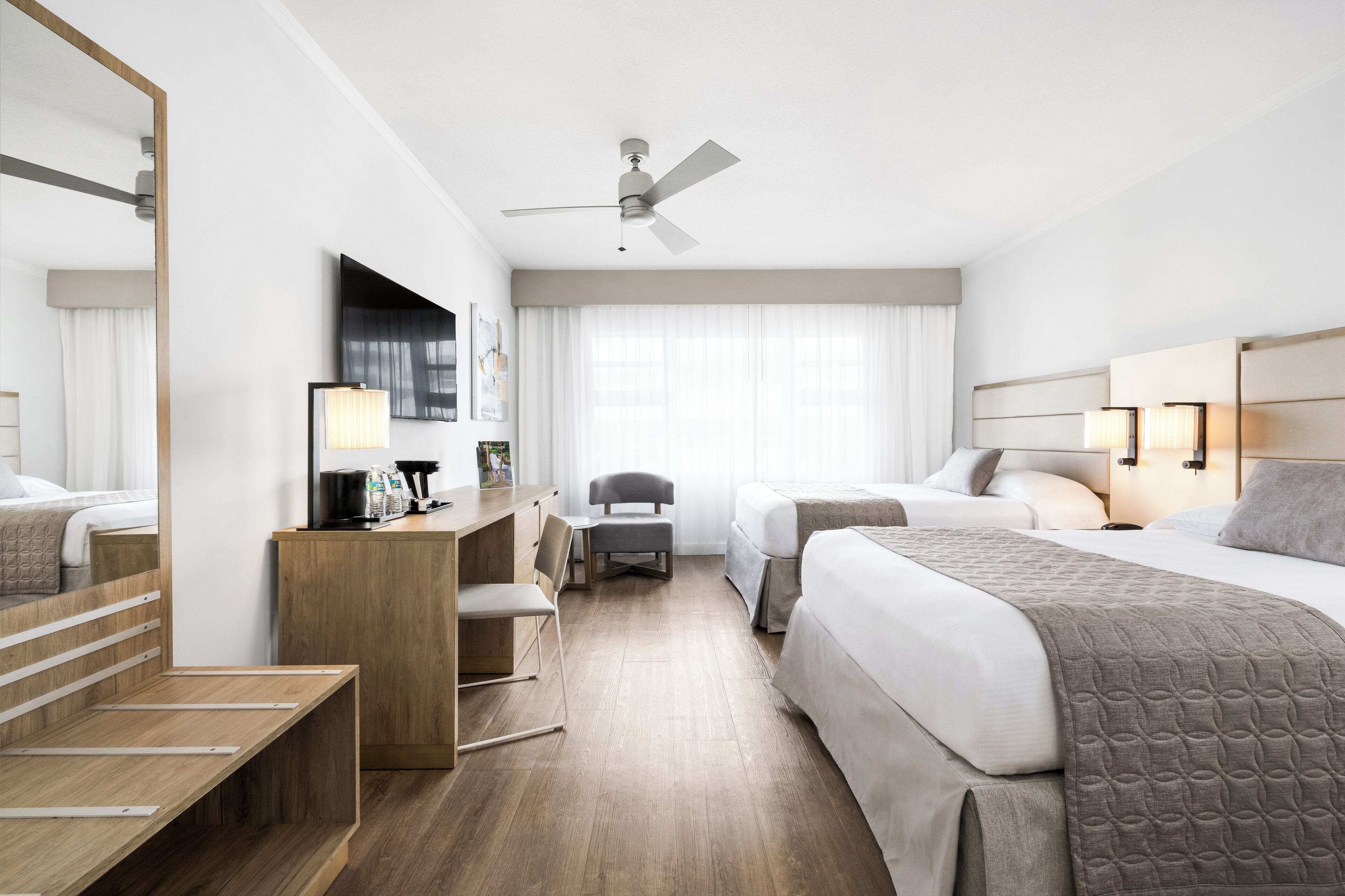 A modern hotel room with a large bed, a desk with a chair, a small table, a ceiling fan, and a large window with curtains.