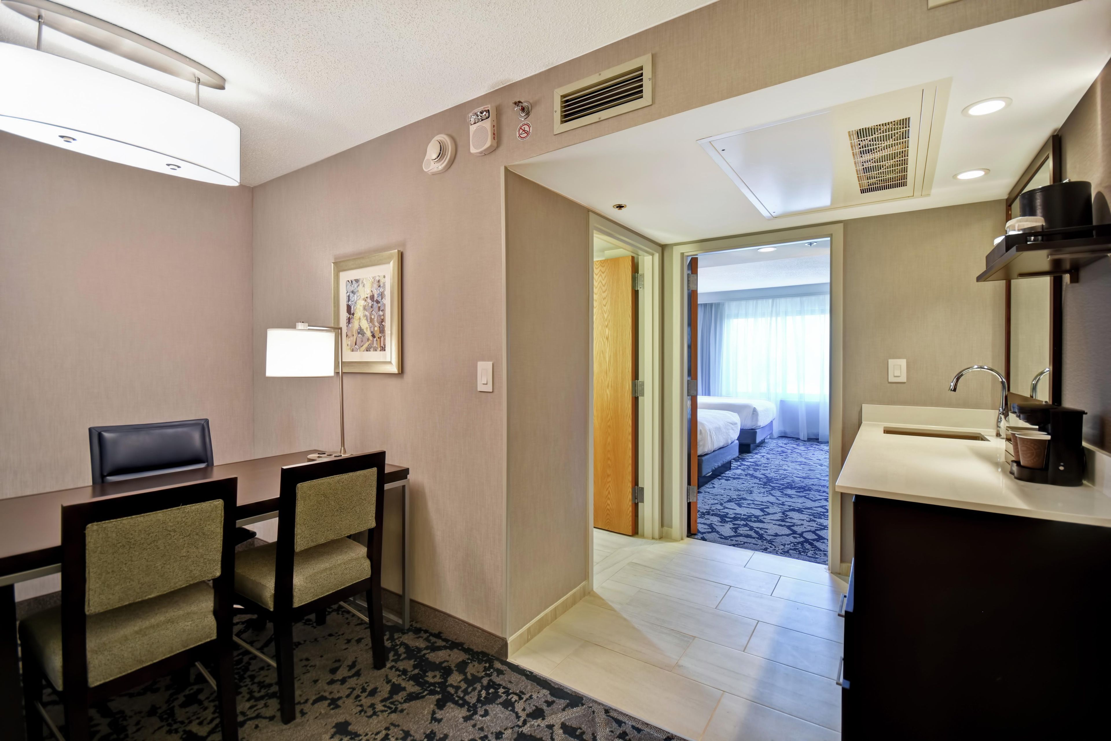 Habitación de hotel con escritorio, sillas, cama, baño con lavamanos y espejo, y un pasillo que da a otra habitación.
