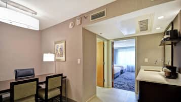 Habitación de hotel con escritorio, sillas, cama, baño con lavamanos y espejo, y un pasillo que da a otra habitación.