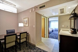 A hotel room with a desk, chairs, a bed, a bathroom with a sink and mirror, and a hallway leading to another room.