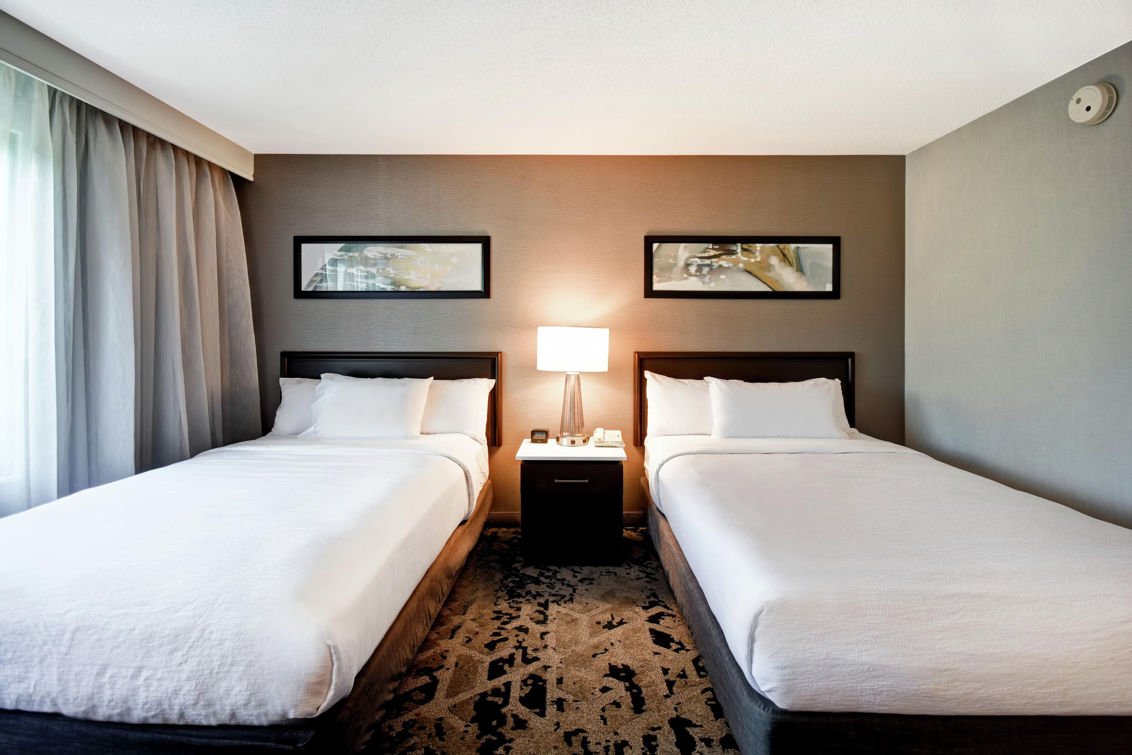 A hotel room with two single beds, a nightstand with a lamp, and two framed pictures on the wall.