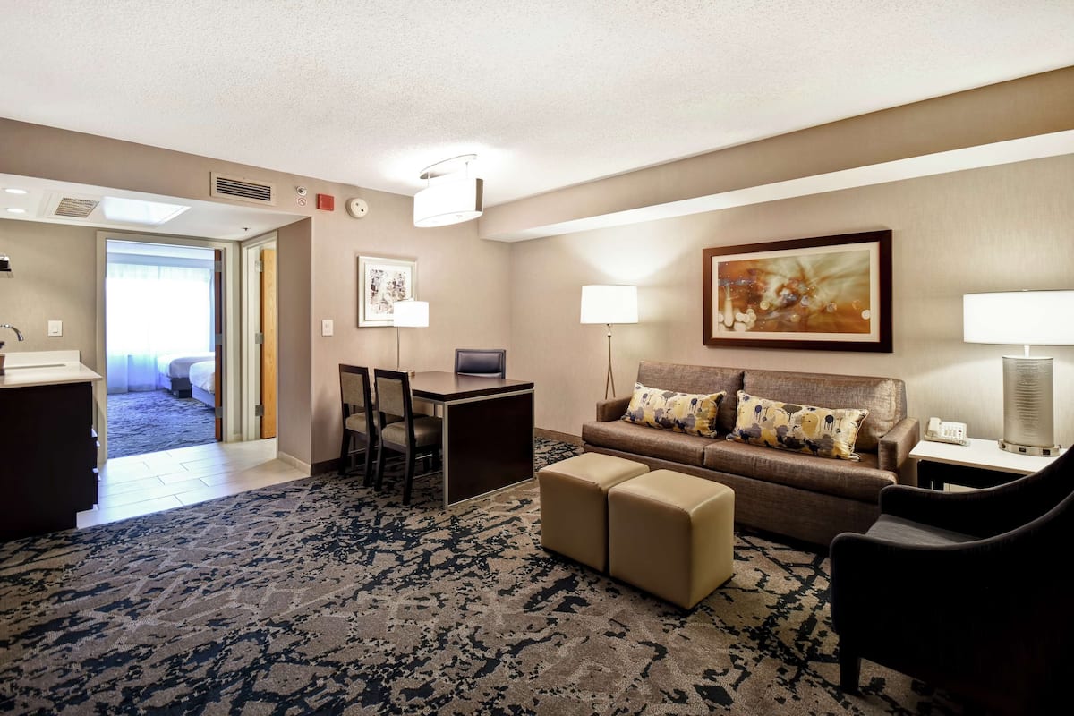 A hotel room with a sofa, ottoman, desk, chair, and artwork on the wall.