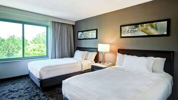 A hotel room with two beds, a large window, a lamp, and two framed pictures on the wall.