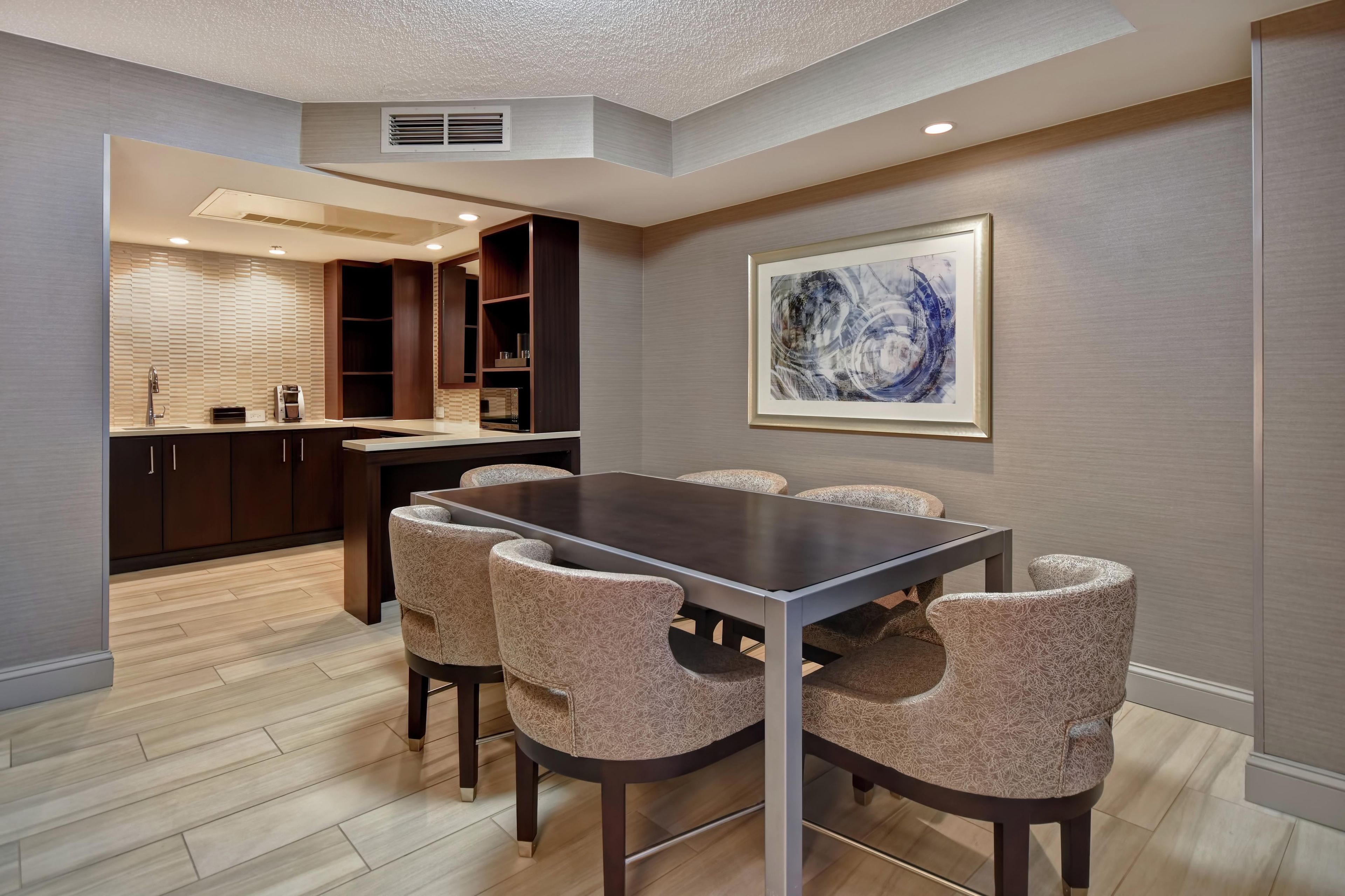 Un espace repas moderne avec une table rectangulaire et six chaises, une cuisine en arrière-plan et un tableau abstrait encadré accroché au mur.