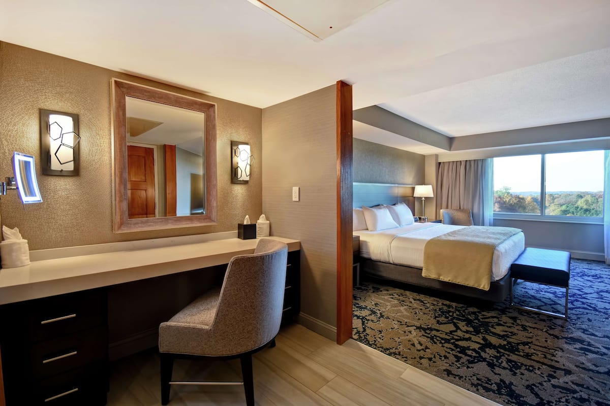 A hotel room with a bed, a desk with a chair, a large mirror, and a view of the outdoors through the window.