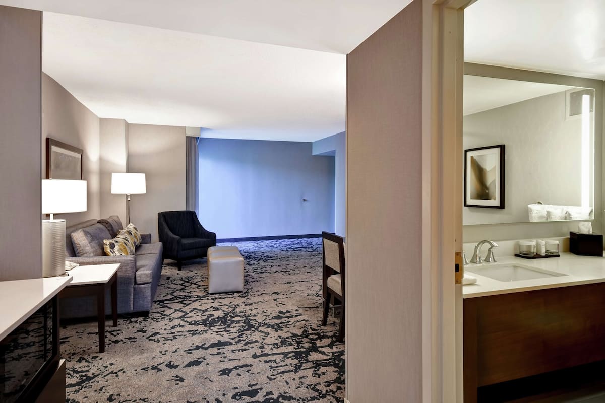 A hotel room with a sofa, armchair, ottoman, and a large window.
