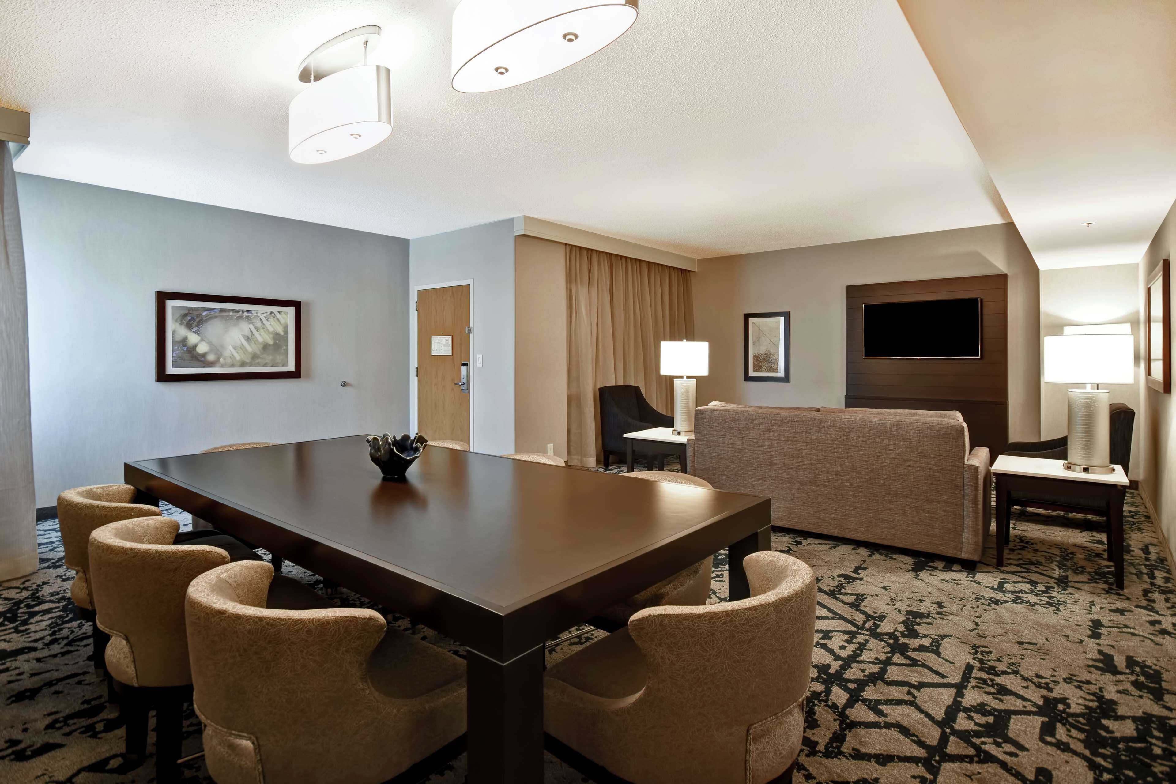 Habitación de hotel con una gran mesa de madera oscura, sillas tapizadas en color beige, un sofá, un televisor montado en la pared y una alfombra estampada.