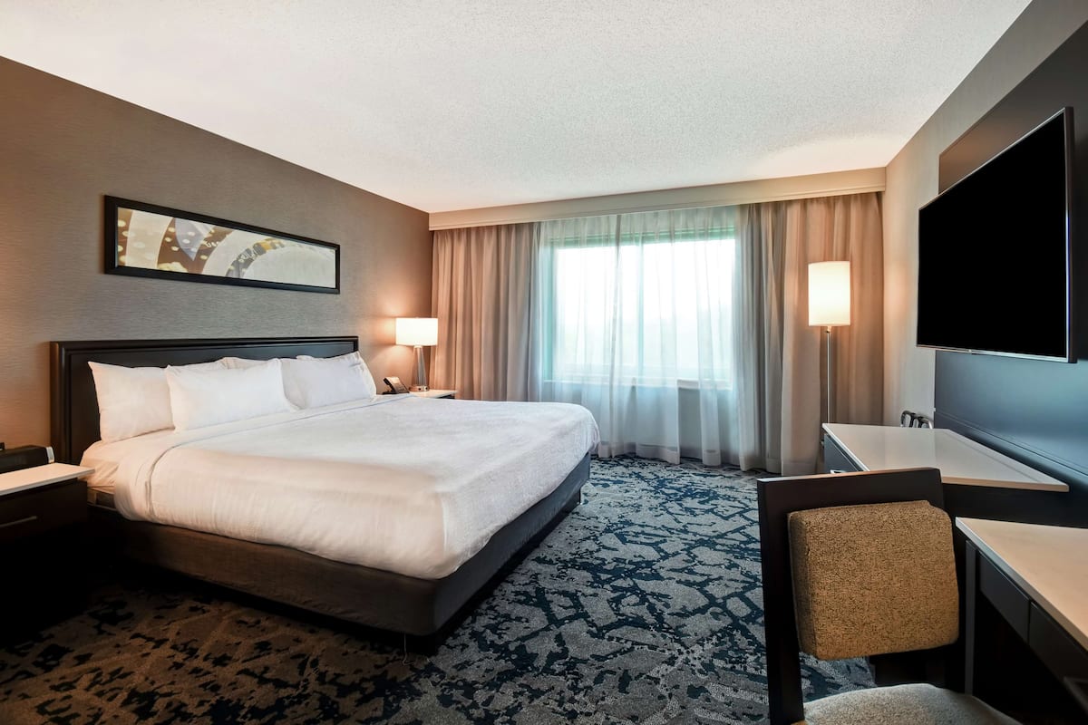 A hotel room with a large bed, a desk with a chair, a flat-screen TV, and a window with curtains.