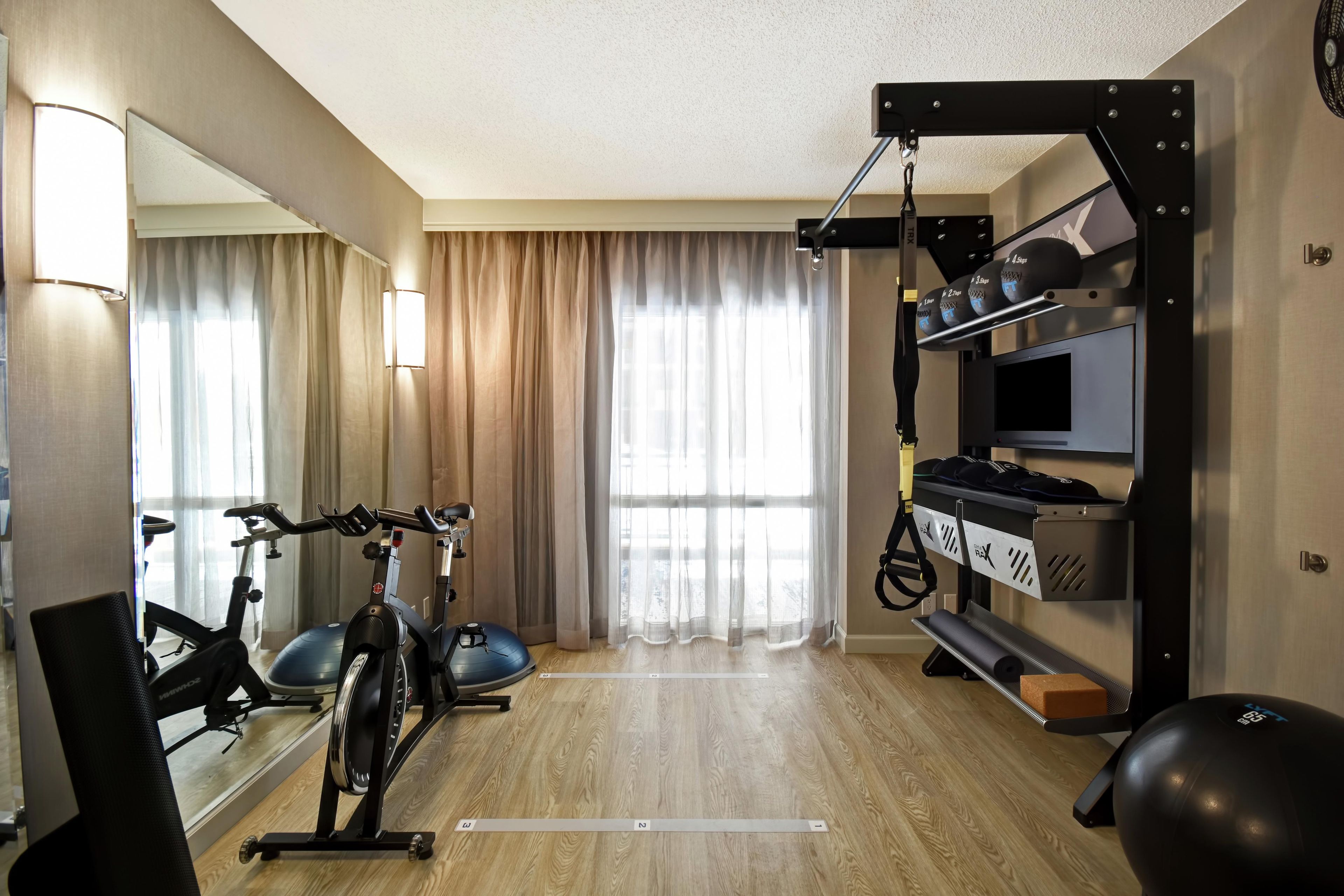 Une salle de sport à domicile équipée de matériel d’exercice, d’un téléviseur et de grandes fenêtres avec des rideaux.