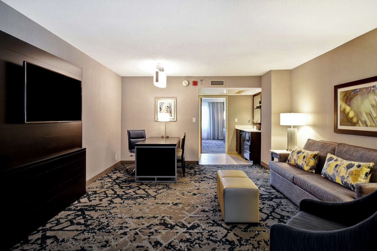 A hotel room with a flat-screen TV, a sofa with patterned cushions, a dining area with a table and chairs, and a patterned carpet.